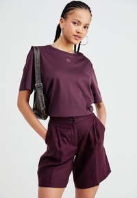 Maroon short-sleeve t-shirt with a small logo detail, paired with high-waisted maroon shorts, featuring pleats and side pockets. Silver chain earrings.