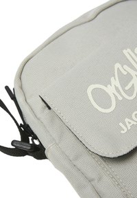 Bolso de bandolera gris de tela con una superficie texturizada y cierre de solapa, que presenta un logo blanco y acentos de hardware negro. Incluye una correa ajustable.