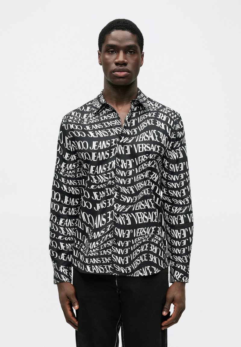 Melnā un baltā krāsā garām piedurknēm krekls ar atkārtotu tekstu, kurā redzami vārdi "VERSACE" un "JEANS." Ar pogāmu apkakli, brīvu piegriezumu, gludu audumu.