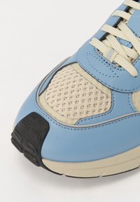 Chaussure de sport bleu clair avec un panneau en maille, des accents beiges, une semelle en caoutchouc texturée et des lacets beiges, avec des détails cousus.