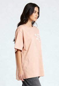 T-shirt en coton oversized rose clair avec des manches courtes, présentant un imprimé graphique blanc sur le devant. Coupe décontractée, texture douce.