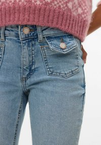 Svijetloplave traperice uskog kroja, s bakrenim kopčama, prednjim džepovima i blagim izblijedjenjem teksture tkanine.