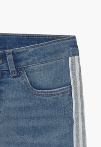 Pantalones vaqueros de mezclilla con cintura de altura media, tela azul, que cuentan con una franja lateral blanca con acentos brillantes y un bolsillo delantero.