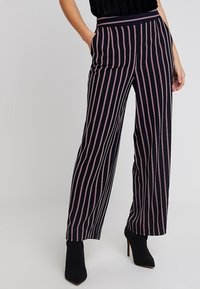 Pantalones de pierna ancha a rayas negras y rosas con una textura suave, cintura alta y líneas verticales que acentúan la figura. Llevados con tacones negros.