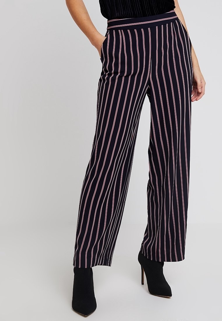 Pantalones de pierna ancha a rayas negras y rosas con una textura suave, cintura alta y líneas verticales que acentúan la figura. Llevados con tacones negros.