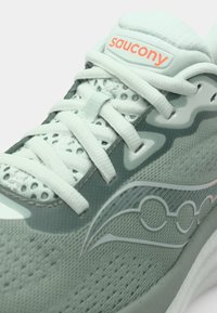 Zapatilla deportiva verde claro con tela de malla, cordones blancos y suela texturizada. Logo destacable y patrón de olas en el lado.