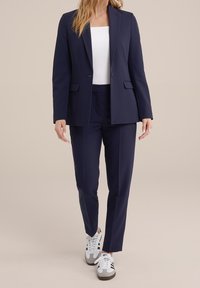 Námořnický modrý blejzr se strukturovaným designem a dvěma předními kapsami, doplněný o odpovídající kalhoty na míru a bílý top. Celý vzhled dokončují pohodlné tenisky.