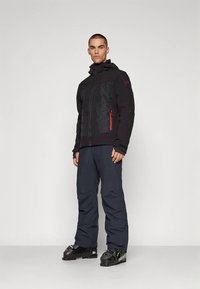 Black insulated jacket with zippered pockets and red accents, paired with dark pants and ski boots, standing against a neutral background.