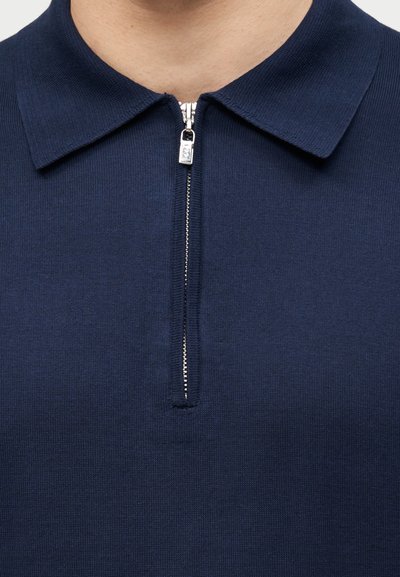 Κοντινό πλάνο ενός πλεκτού πουκάμισου ναυτικού μπλε με ασημί φερμουάρ μερικώς ανοιχτό κάτω από μια μυτερή γιακά.