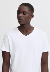 Persoon met een effen wit T-shirt met V-hals kijkt omlaag en opzij tegen een effen lichte achtergrond.