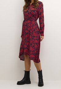 Donna con un abito midi blu navy con un motivo floreale rosso e stivali neri massicci alla caviglia, in piedi di fronte a uno sfondo bianco semplice.