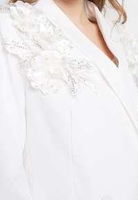 Biała marynarka z kwiatowymi zdobieniami i koralikowymi detalami na ramieniu. Posiada klapki o wycięciu oraz gładką teksturę materiału.