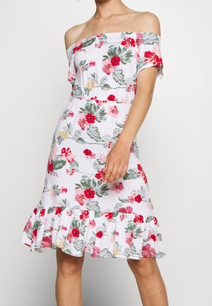 Kobieta w białej sukience odsłaniającej ramiona z czerwonym i różowym kwiatowym wzorem oraz marszczonym dołem, stojąca na jednolitym jasnym tle.