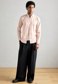 Ανοιχτό ροζ πουκάμισο με κουμπιά, σε συνδυασμό με φαρδιά μαύρα παντελόνια και μαύρα παπούτσια, στέκεται σε ξύλινο δάπεδο μπροστά από φωτεινό φόντο.