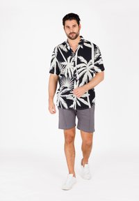 Maglietta nera a maniche corte con stampa di palme bianche, abbinata a pantaloni corti grigi e sneakers bianche. Il modello è in piedi su uno sfondo bianco.