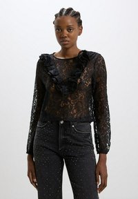 Blusa nera in pizzo con maniche lunghe, scollatura arricciata e motivi floreali. Abbinata a jeans neri decorati con borchie argentate.