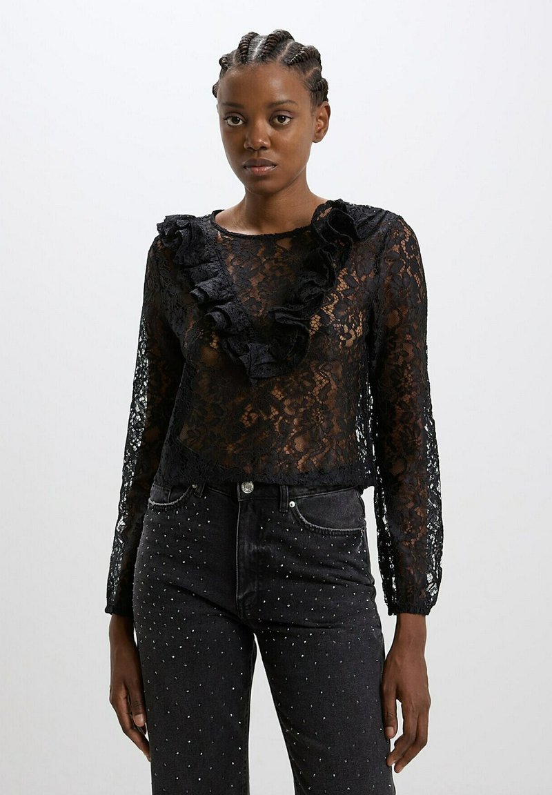 Blusa nera in pizzo con maniche lunghe, scollatura arricciata e motivi floreali. Abbinata a jeans neri decorati con borchie argentate.