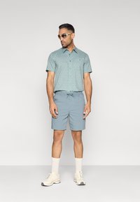 Ανοιχτό μπλε κοντομάνικο πουκάμισο με σχέδιο και γιακά, συνδυασμένο με ανοιχτό μπλε σορτς. Αξεσουάρ περιλαμβάνουν λευκά αθλητικά παπούτσια και γυαλιά ηλίου.