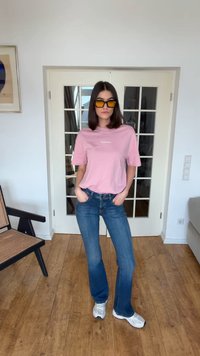 Γυναίκα με σκούρα μαλλιά μέχρι τους ώμους, ντυμένη με ροζ μπλουζάκι, μπλε τζιν, λευκά αθλητικά παπούτσια και γυαλιά ηλίου με κεχριμπαρένιο φακό, στέκεται σε δωμάτιο με ξύλινο πάτωμα.