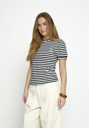 Kobieta z długimi włosami, ubrana w krótki rękaw w czarno-białe paski i białe spodnie z wysokim stanem, stojąca na tle jednolitego białego tła.