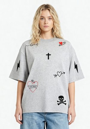 Blond vrouw draagt een grijze oversized T-shirt met zwart- en roodkleurige grafische prints, waaronder een kruis, harten, bliksemschichten, schedel en de tekst "Love Hurts".