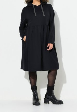 Femme portant une robe à capuche noire à manches longues avec une taille froncée, des collants noirs et des bottines noires à talons, debout sur un fond blanc.