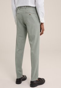 Šviesiai žali, tekstūruoti prilipinti kelnių modeliai su siauru siluetu, dviem galinėmis kišenėmis ir rudos spalvos mygtukais, derinami su juodais batais.