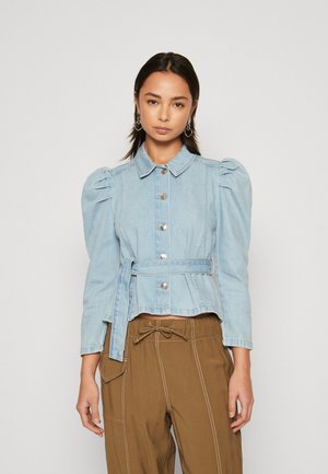 Gai zilas denim jaka ar pūkainām piedurknēm, piegulošu vidukli, sudraba pogu aizdari un atbilstošu jostu, apvienota ar brūnajiem biksēm.