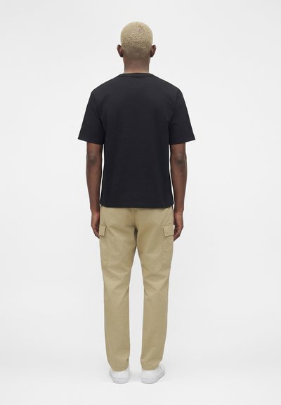 Άνδρας με κοντά ξανθά μαλλιά, φορώντας μαύρο μπλουζάκι, μπεζ παντελόνια cargo και λευκά αθλητικά παπούτσια, στέκεται με την πλάτη σε απλό φόντο.