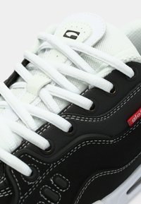 Baskets en noir et blanc avec une tige en cuir texturé, des lacets blancs, des coutures visibles et un accent de logo rouge sur le côté.