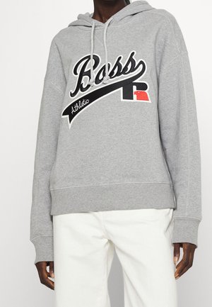 Personne portant un sweat à capuche gris avec le logo "Boss Athletic" en noir et blanc, associé à un pantalon blanc, visible des épaules aux hanches.