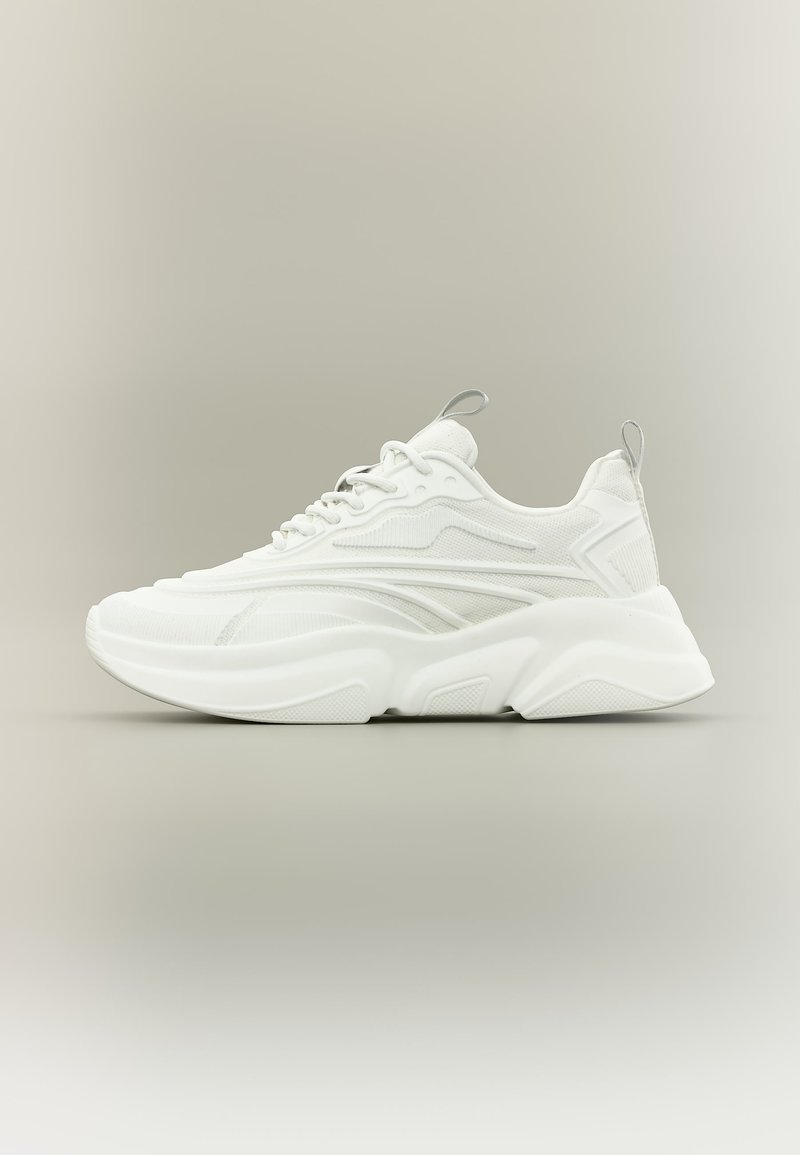 Baskets de sport blanches avec semelle épaisse, dessus en mesh et synthétique, fermeture à lacets, et languettes de tirage sur la langue et le talon, vue de côté sur un fond neutre.