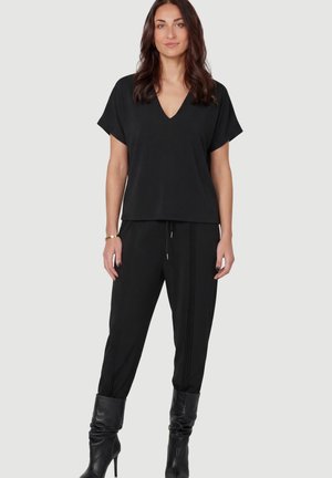 Vrouw draagt een zwarte V-hals korte mouw top, zwarte broek met trekkoord en verticale strepen, en zwarte hoge hakken, staand tegen een effen achtergrond.