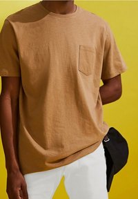 Personne portant un t-shirt beige à manches courtes avec une poche et un pantalon blanc, tenant une casquette noire derrière le dos sur un fond jaune.
