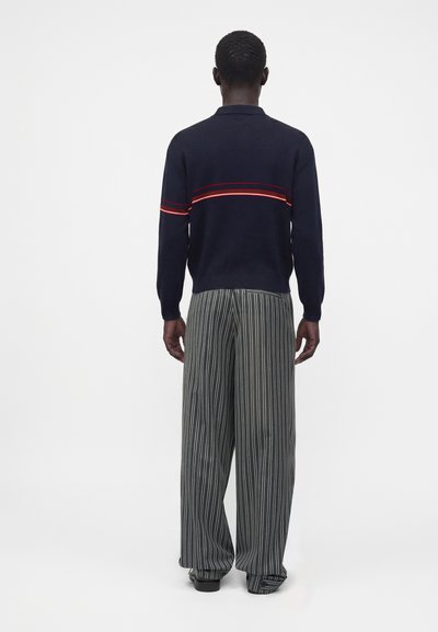 Homme de dos portant un pull bleu marine à rayures rouges et un pantalon ample rayé verticalement noir et blanc sur un fond uni.