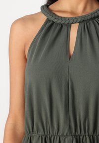 Vestido sem mangas de cor verde oliva, com decote trançado, detalhe frontal em forma de olho-chave e cintura franzida para um ajuste descontraído.