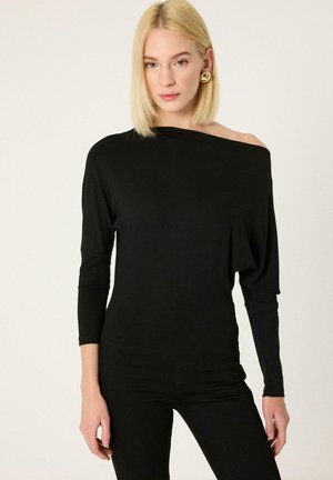 Donna bionda con top nero scollato sulle spalle a maniche lunghe e pantaloni neri, accessoriata con grandi orecchini dorati, su sfondo uniforme.