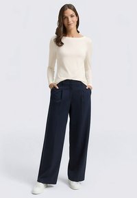 Cremefarbenes Langarmshirt kombiniert mit marineblauen, weit geschnittenen Hosen. Die Hosen sind mit Falten und Seitentaschen ausgestattet. Das Modell trägt weiße Turnschuhe.