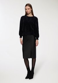 Kobieta stoi w czarnym, puszystym swetrze, czarnej spódnicy do kolan z rozcięciem z przodu, przezroczystych rajstopach i czarnych, spiczastych botkach za kostkę na białym tle.