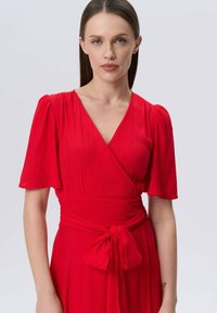 Rotes Wickelkleid mit kurzen, gepufften Ärmeln, einem tiefen V-Ausschnitt und einem Stoffband in der Taille; strukturiertes Material sorgt für visuelles Interesse.