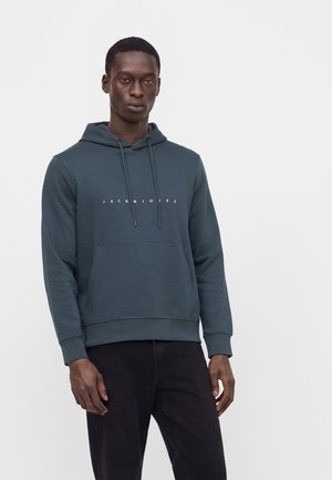 Hombre con una sudadera con capucha azul oscuro con el texto "JACK & JONES" y pantalones negros, de pie frente a un fondo liso gris claro.