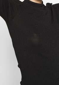 Haut noir ajusté en tissu texturé avec une légère étincelle. Col montant et manches courtes, avec un détail froncé à l'épaule.