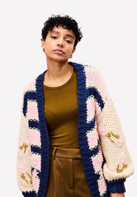 Frau mit kurzen, lockigen Haaren, die einen grob gestrickten Cardigan mit Blumen- und geometrischen Mustern über einem braunen Shirt und passenden Hosen trägt.