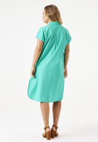 Türkises Hemdkleid mit kurzen aufgerollten Ärmeln, einer Rückenfalte und einem abgerundeten Saum, kombiniert mit braunen High-Heels-Sandalen.