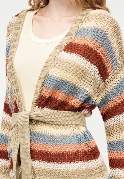 Πλεκτή ζακέτα με οριζόντιες ρίγες σε μπεζ, μπλε, σκουροκόκκινο, λευκό και ροδακινί, δεμένη στη μέση πάνω από μια εκρού μπλούζα με στρογγυλή λαιμόκοψη.