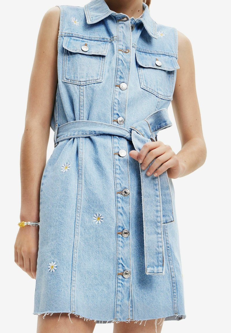 Robe sans manches en denim bleu clair avec boutons sur le devant, ceinture nouée à la taille, poches poitrine et petites marguerites blanches brodées.