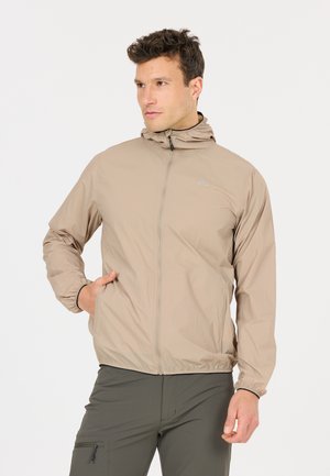 Mann trägt beige Kapuzen-Sweatjacke mit Reißverschluss und einer Hand in der Tasche, steht vor einem schlichten hellen Hintergrund.