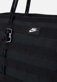 Mustmust musta kott, mis on mustast Nike'ist, koos tekstureeritud kangaga, punutud rihmadega ja väikese logoga plaastriga. Sisaldab veebimisdetaili hoidmiseks või kinnitamiseks.