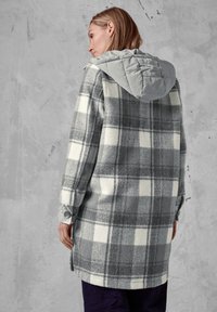 Rutet frakke i grå og hvid fleece med en grå quiltet hætte. Har knaplukning ved manchetterne og sideslidser for fri bevægelse.