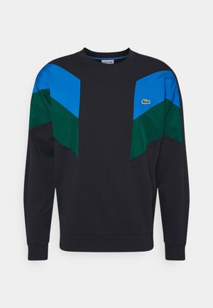 Musta huppari, jossa on rohkeat siniset ja vihreät väri-askelmat harteilla. Ominaisuuksiin kuuluu pyöreä pääntie ja ribbikudotut hihansuut. Lacoste-logo rinnassa.